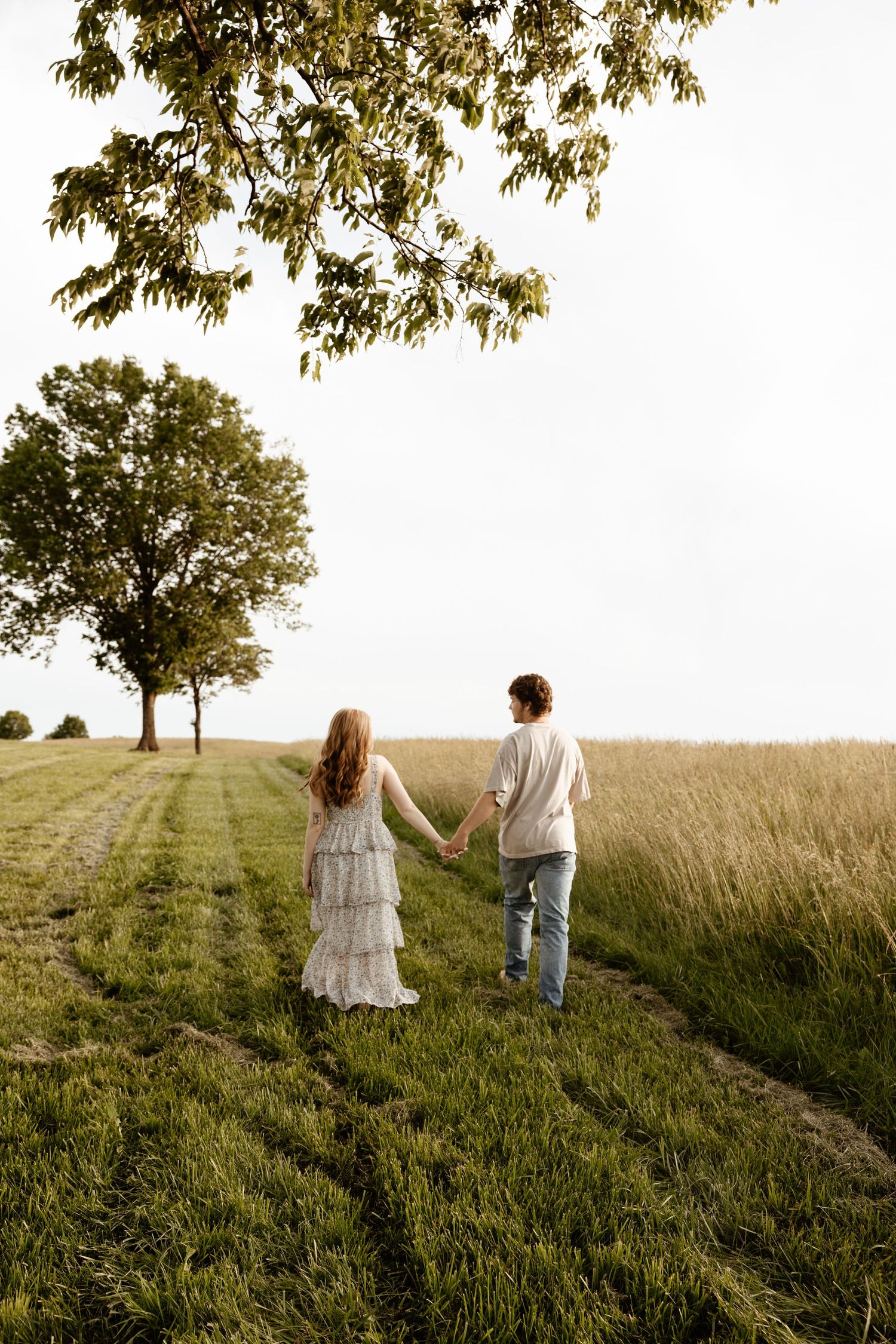 Couple walking hand in hand along a green field path toward trees in soft evening light.