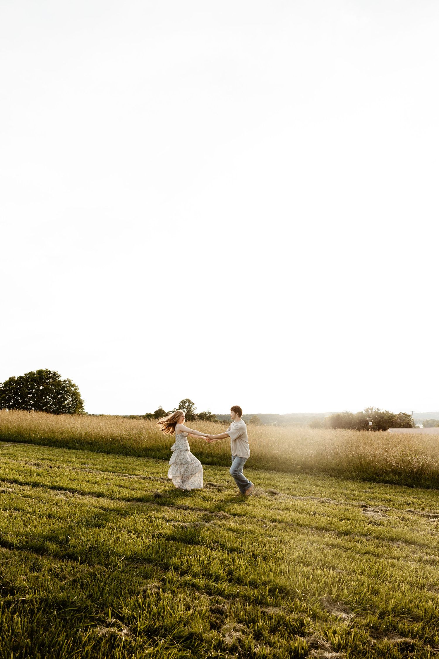 Couple holding hands and running playfully across a green field in bright golden sunset light.
