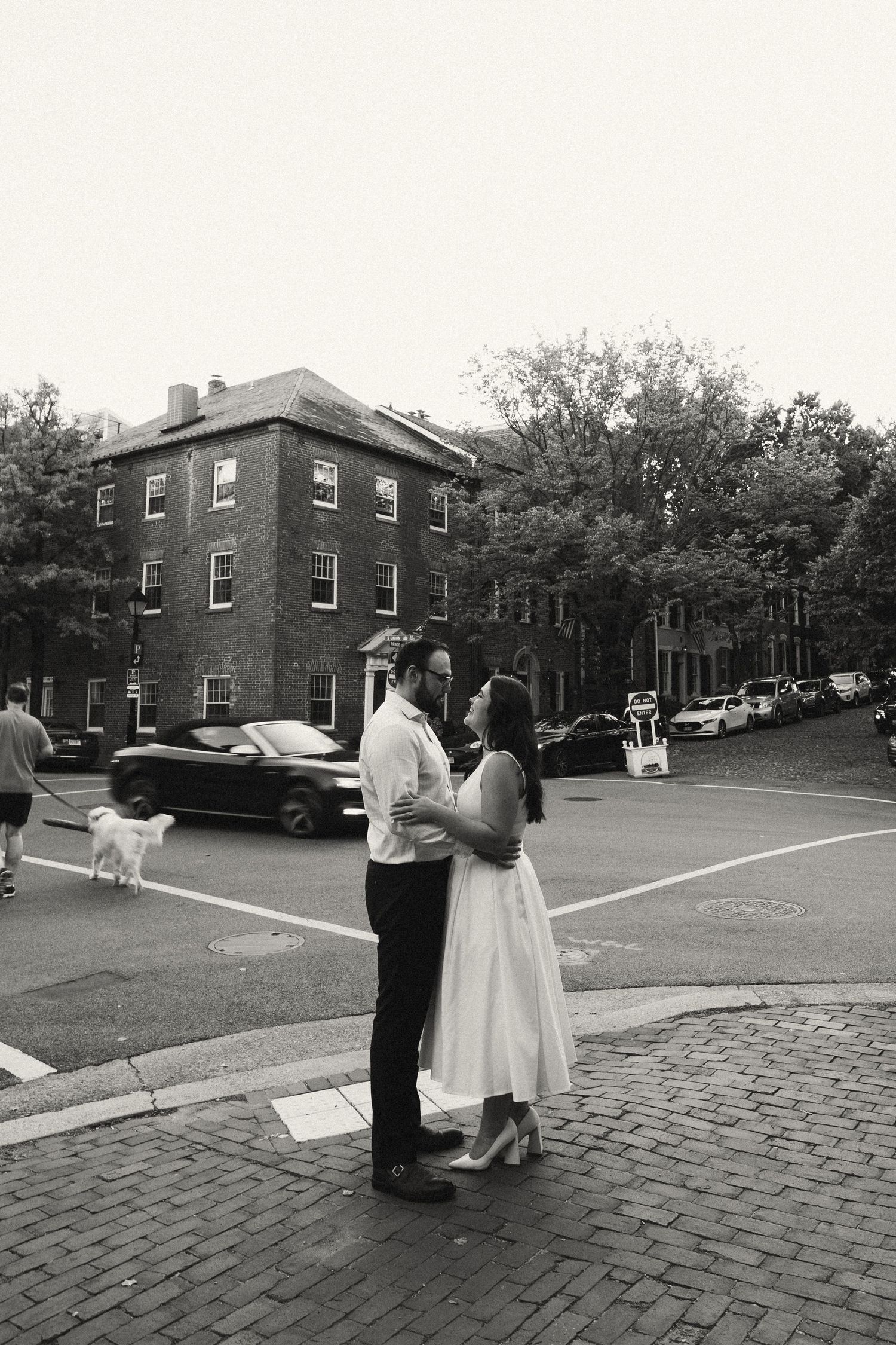 Black and white photo of couple embracing on a cobblestone street corner with historic brick buildings in background.