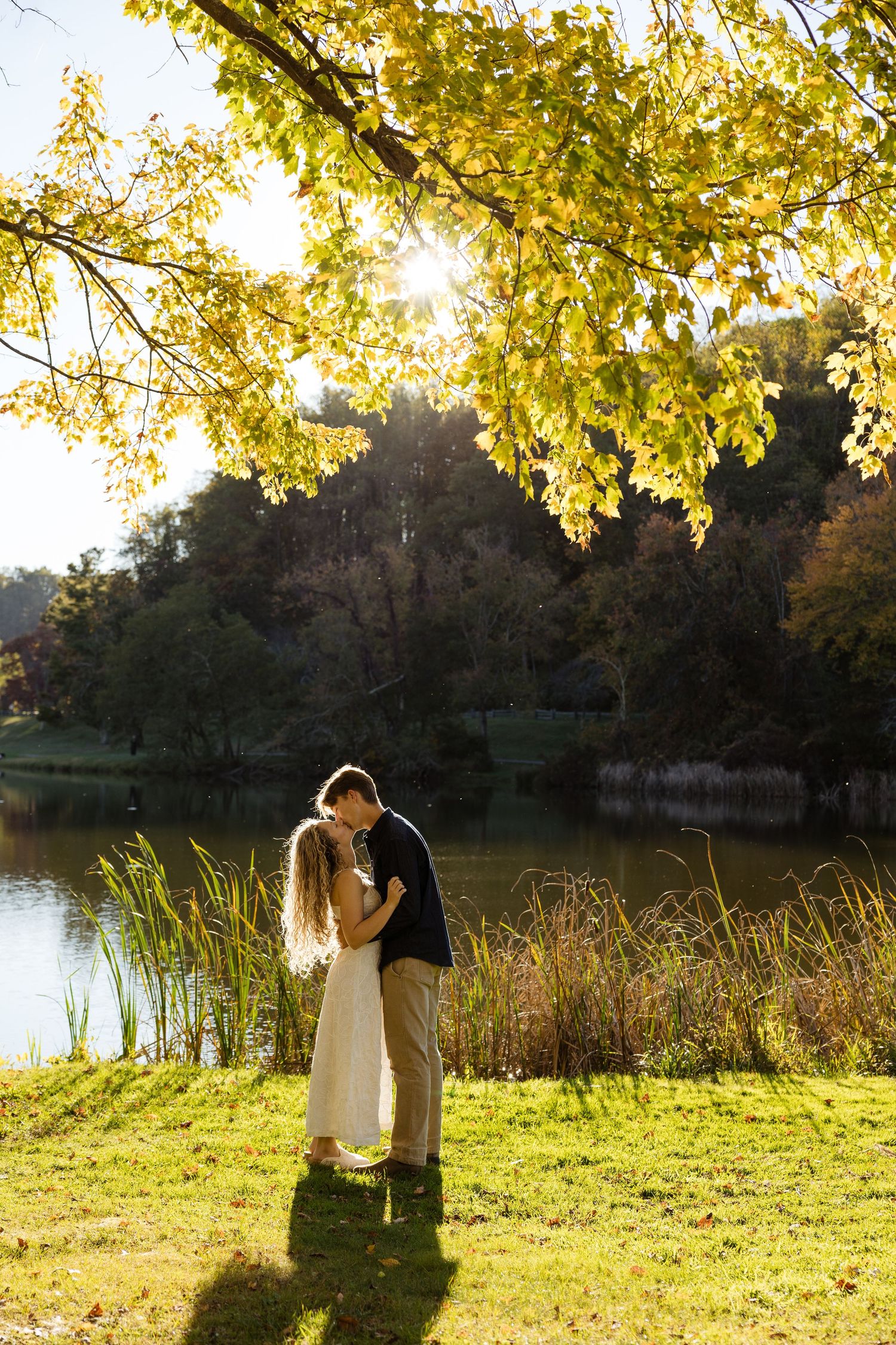 Couple embracing and kissing by a sunlit lake surrounded by autumn foliage in a romantic outdoor setting.