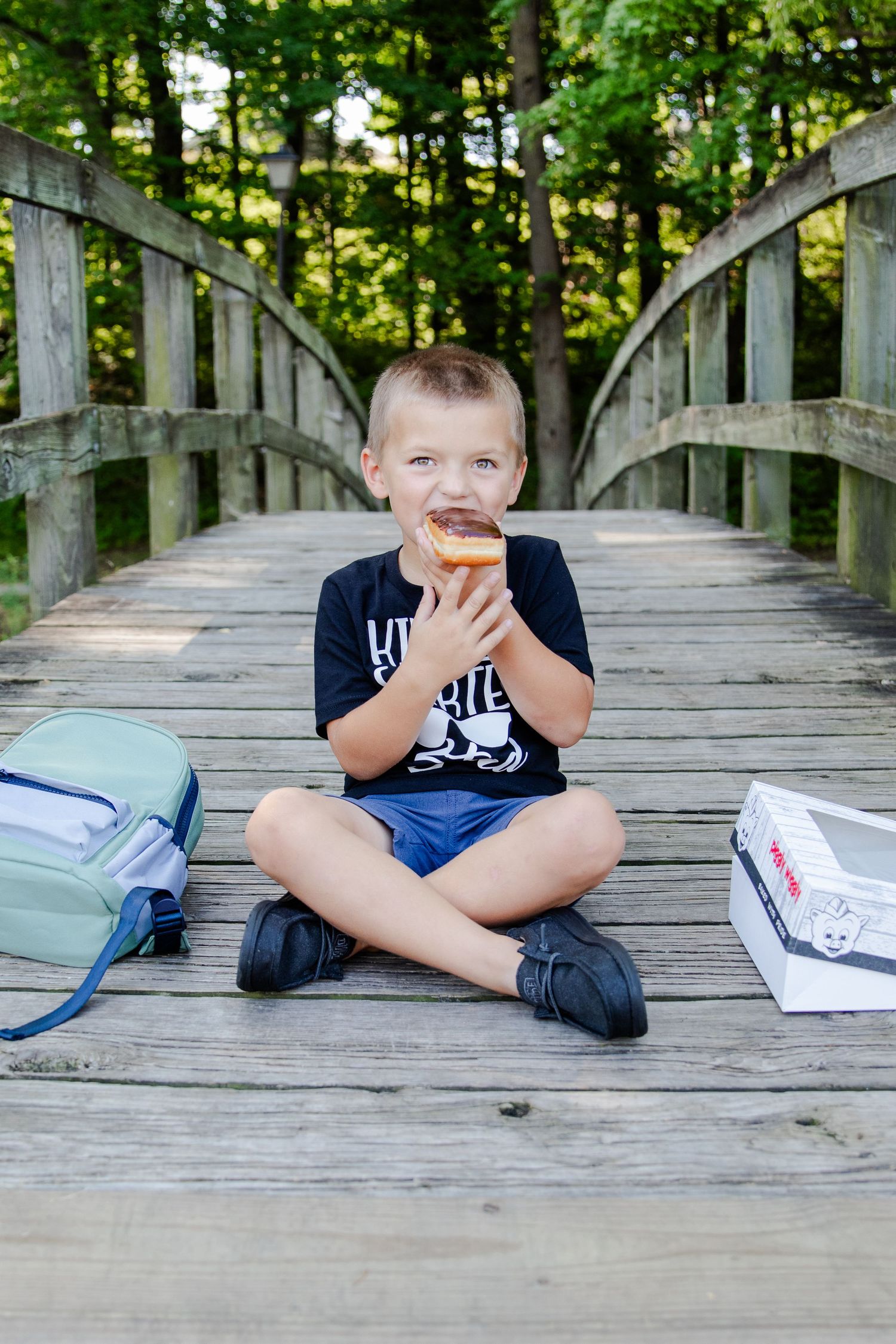 Kindergartener sitting on a bridge with his backpack and eating a donut getting ready for his first day of school.
