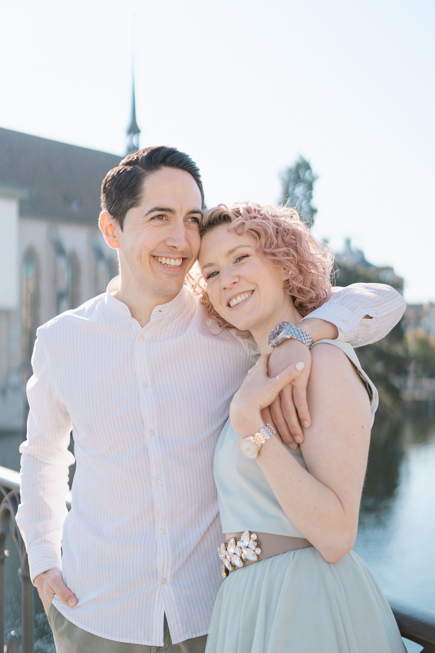 A happy couple embraces on a bridge with a church spire visible in the background.