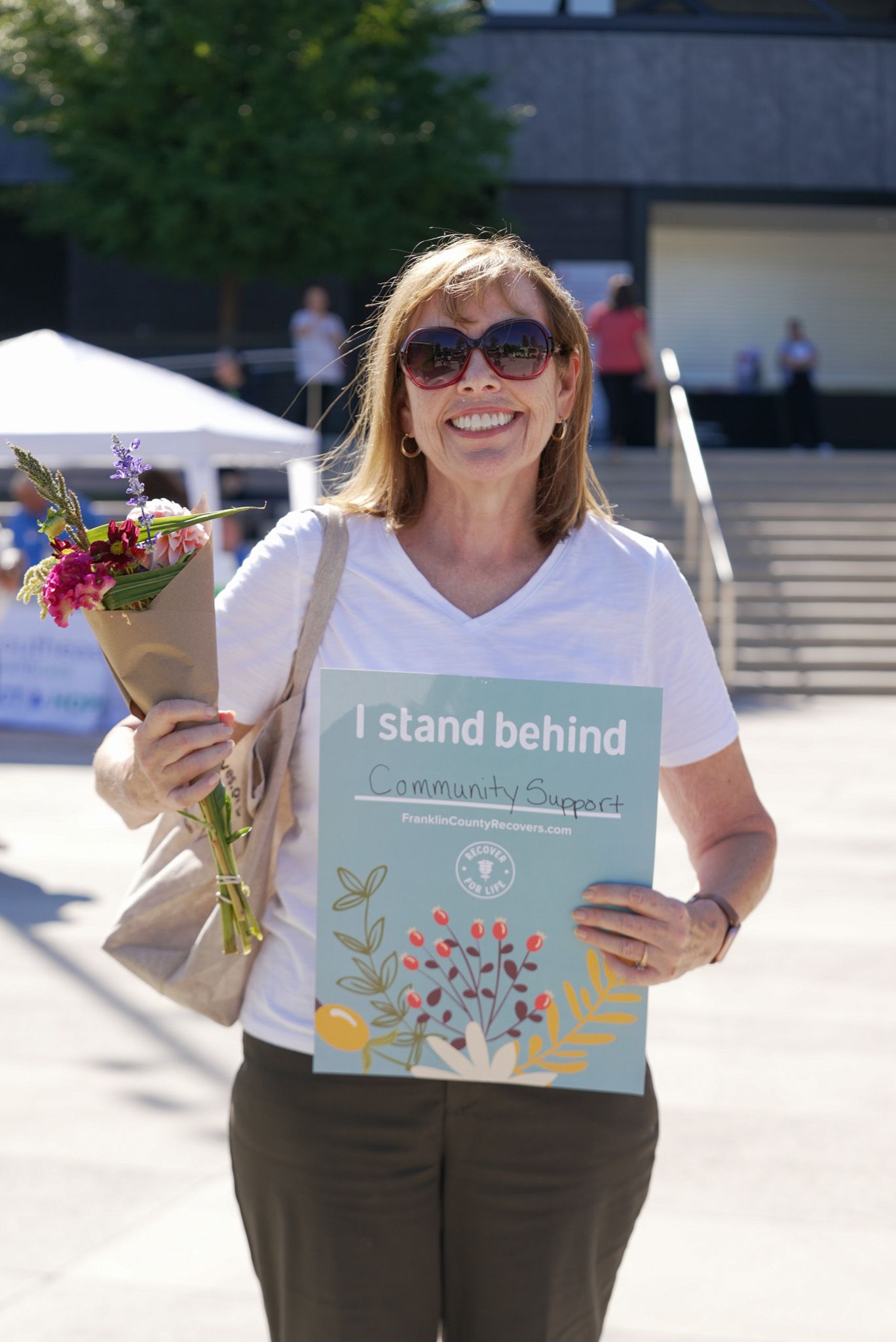 Smiling woman holding flowers and community support sign at an outdoor event.