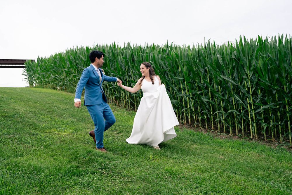 Bride and groom holding hands running along a green grass path beside a tall cornfield on their wedding day.
