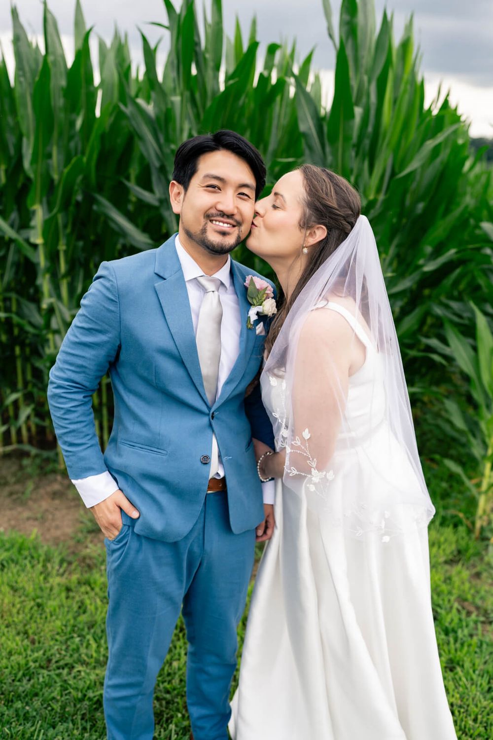 Bride in white gown and veil kisses groom in blue suit cheek outdoors near green cornfield.