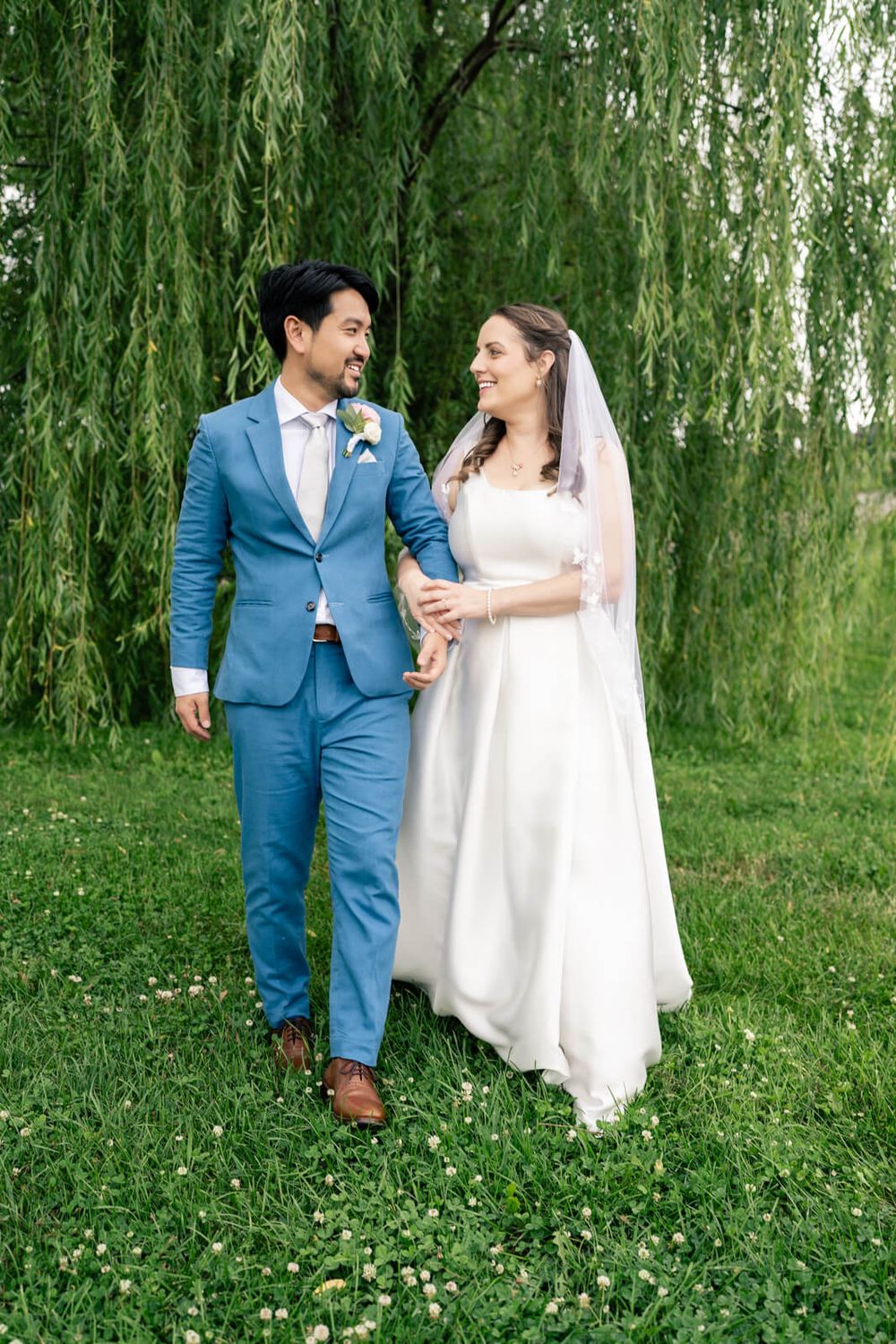 A bride and groom walk hand in hand under a weeping willow tree, smiling at each other on their wedding day.