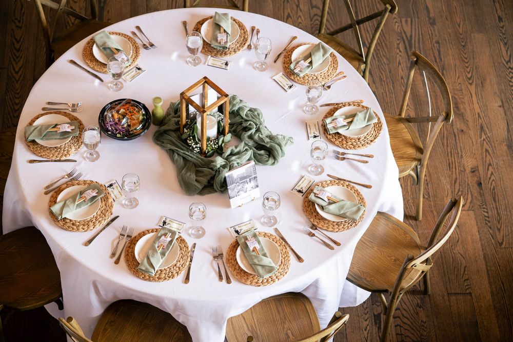 Elegant round wedding reception table with wicker chargers, gold lantern centerpiece, and sage green napkins on white linen.