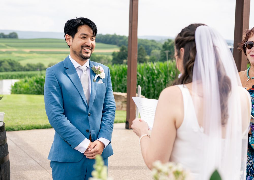 Groom in blue suit smiling at bride during outdoor wedding ceremony with green fields in background.