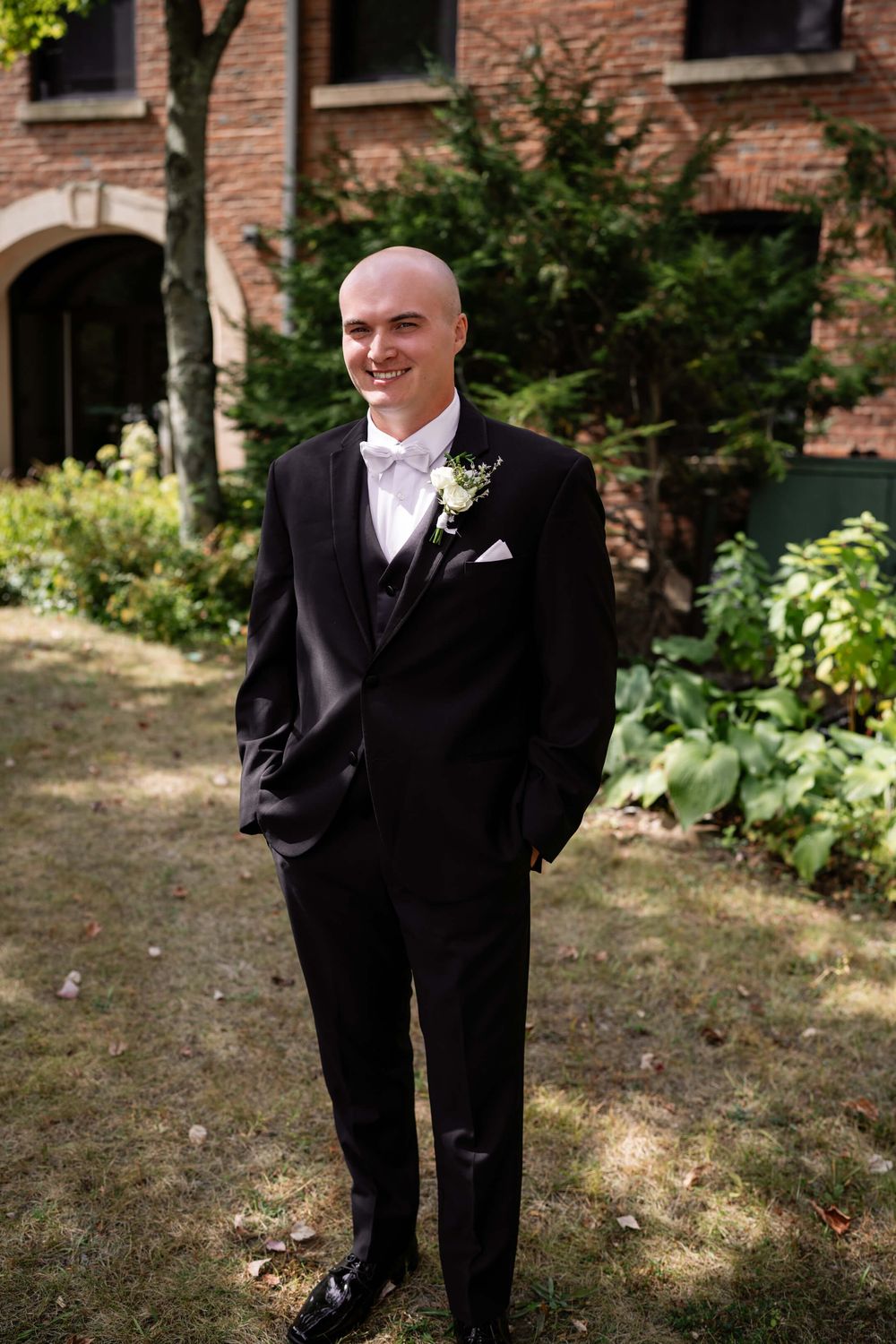 Groomsmen in classic black tuxedos stand together on a lawn with a brick building backdrop.