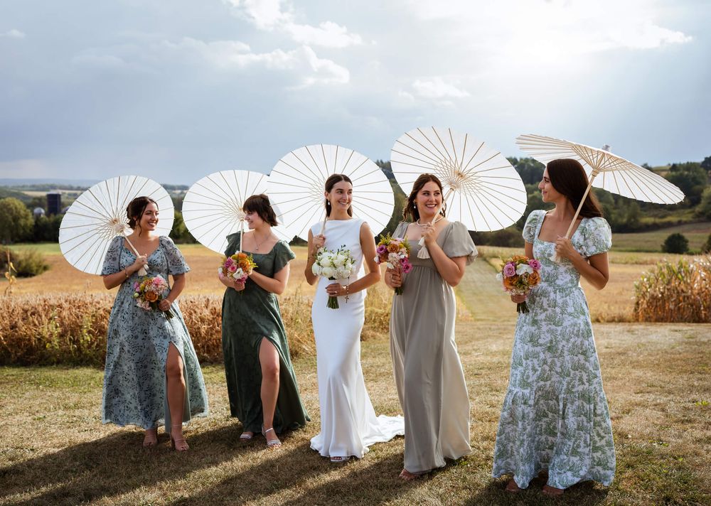 A bridal party poses in a sunny field holding white paper parasols while wearing floral and solid colored dresses.