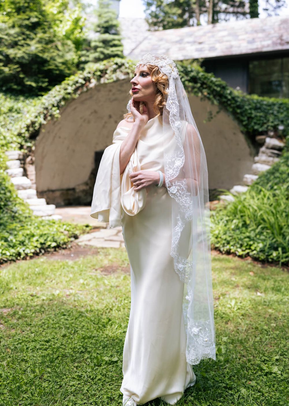Bride in ivory satin gown with lace-trimmed veil and floral crown poses in lush garden near stone archway.