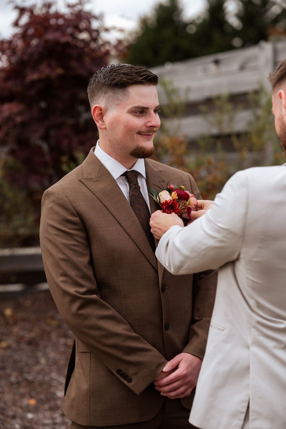 A person in a brown suit receives a boutonniere from someone wearing white in this wedding party preparation moment.