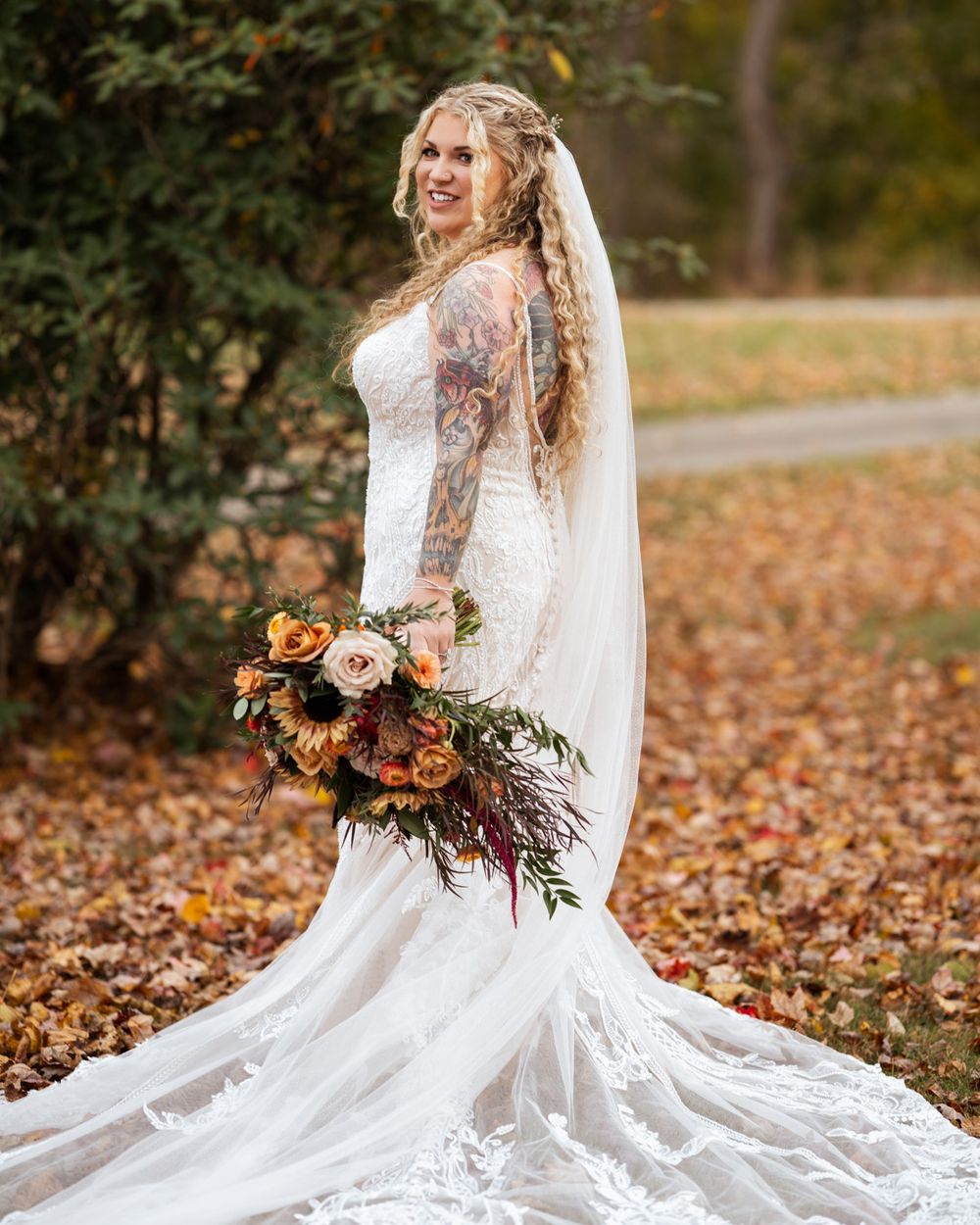 A bride in a white lace gown sits gracefully among autumn leaves holding a vibrant fall-colored bouquet.