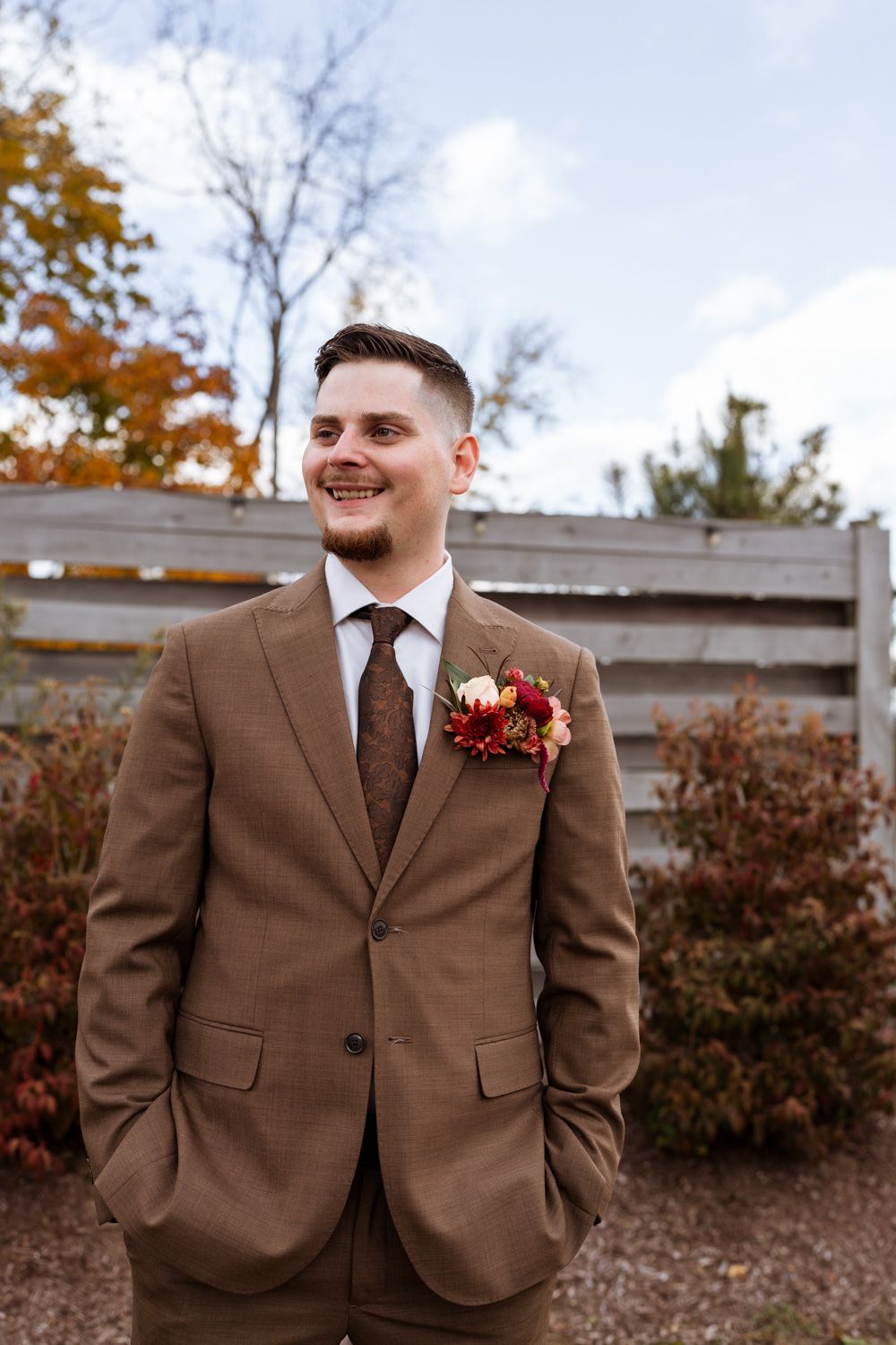 A groom in a brown suit with a fall boutonniere smiles outdoors on an autumn day with fall foliage in background.