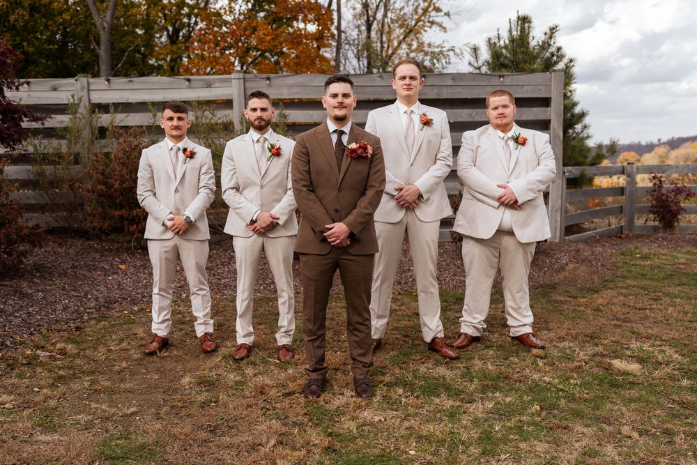 Wedding party in beige suits poses together outdoors during autumn with rustic scenery in the background.