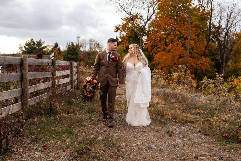 A couple walks together along a rustic wooden fence surrounded by vibrant autumn foliage in a romantic outdoor setting.