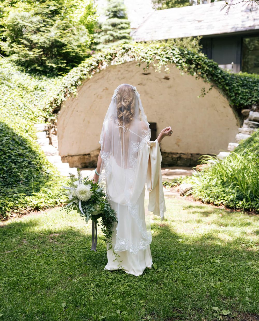 Bride in white gown and flowing veil stands in lush garden beside floral arrangement, facing stone arch backdrop.