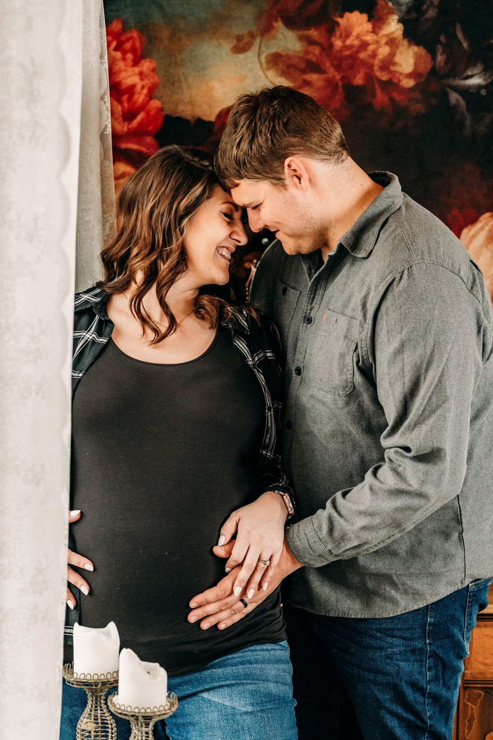 A romantic maternity photoshoot of a couple embracing and laughing in front of a dark floral backdrop.   Creative Photographer, Grand Rapids