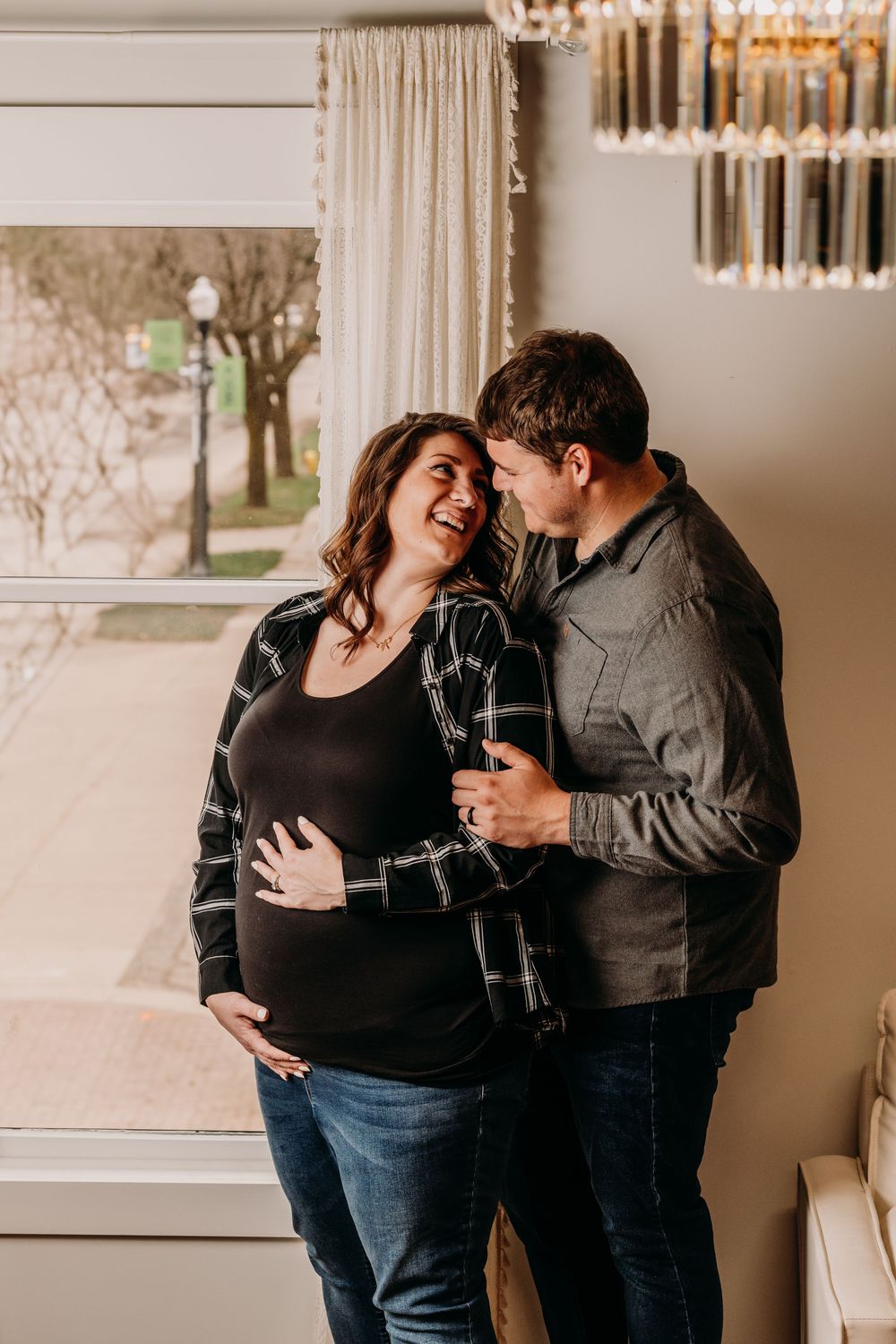An intimate indoor maternity session shows a couple sharing tender moments near a window with soft lighting. Photographer Grand Rapids