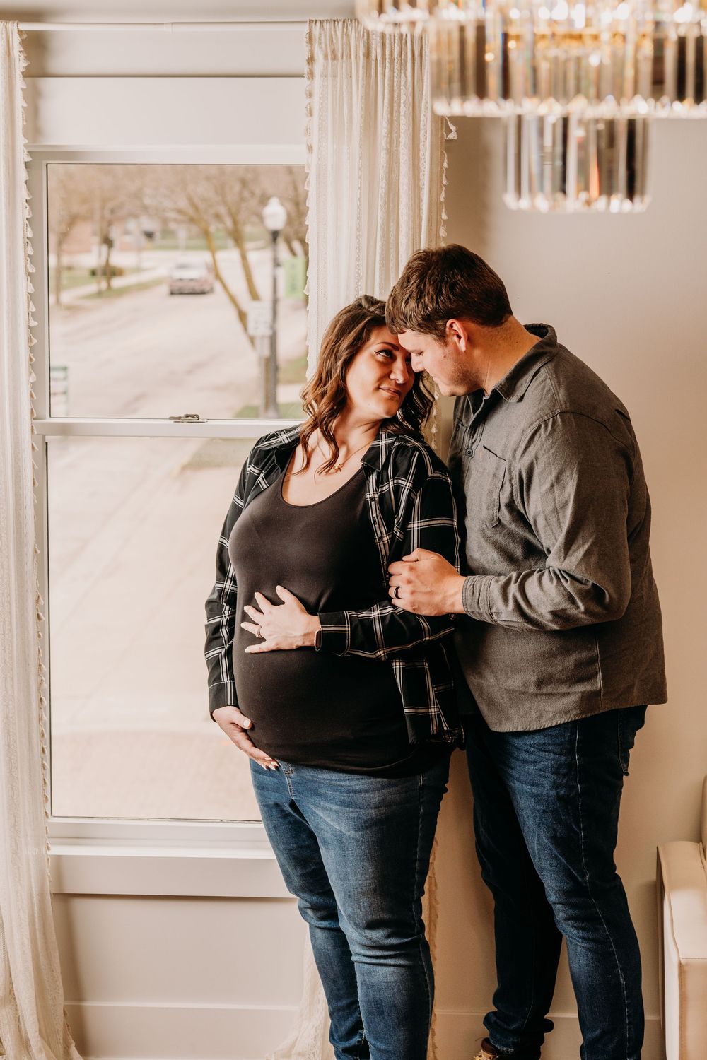 A tender pregnancy portrait of a couple embracing by a window in their home with natural light streaming in. Photography, Grand Rapids MI