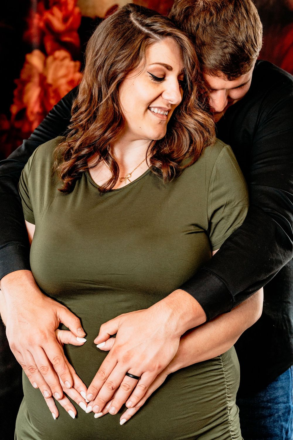 Couple shares intimate moment making heart shape with hands on pregnant woman's belly in olive green dress. Photography Grand Rapids, MI
