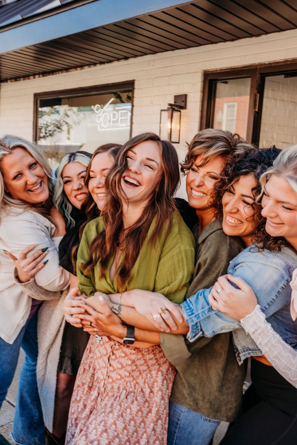 A group of friends laugh together while embracing in casual outfits outside a building on a sunny day.  Headshot Photographer, Grand Rapids