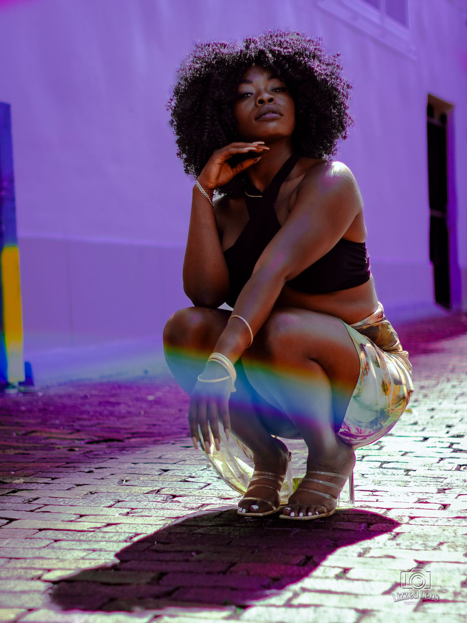 Young Black woman with natural afro crouching against purple wall with rainbow light effect.