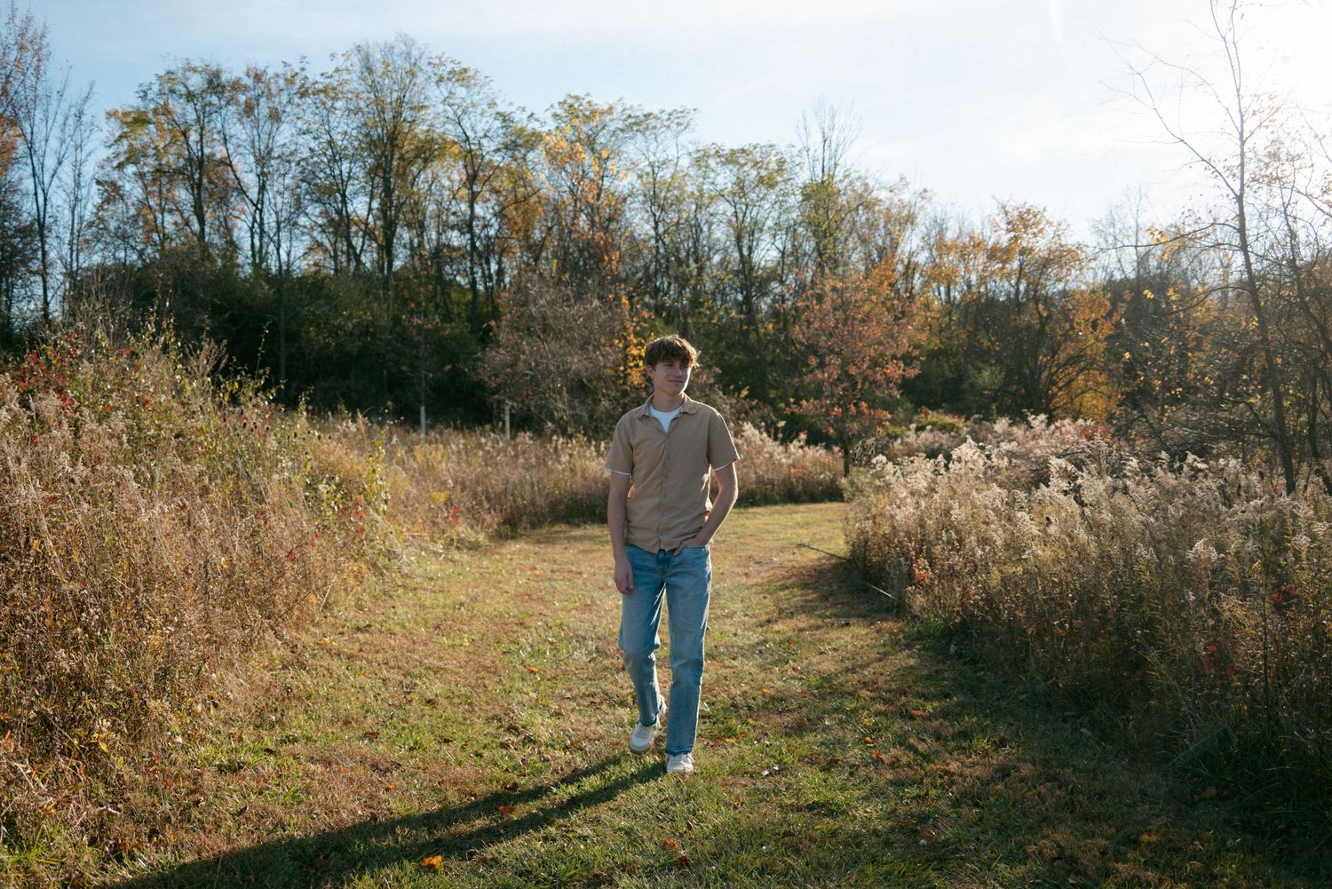 Young man in beige sweater and jeans walking along sunny autumn meadow path with tall dry grasses.