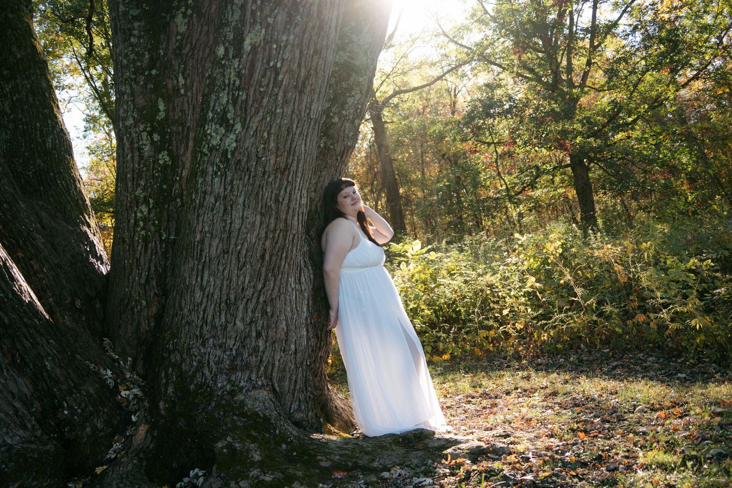 Woman in white gown leans against a large tree in a backlit autumn woodland setting.