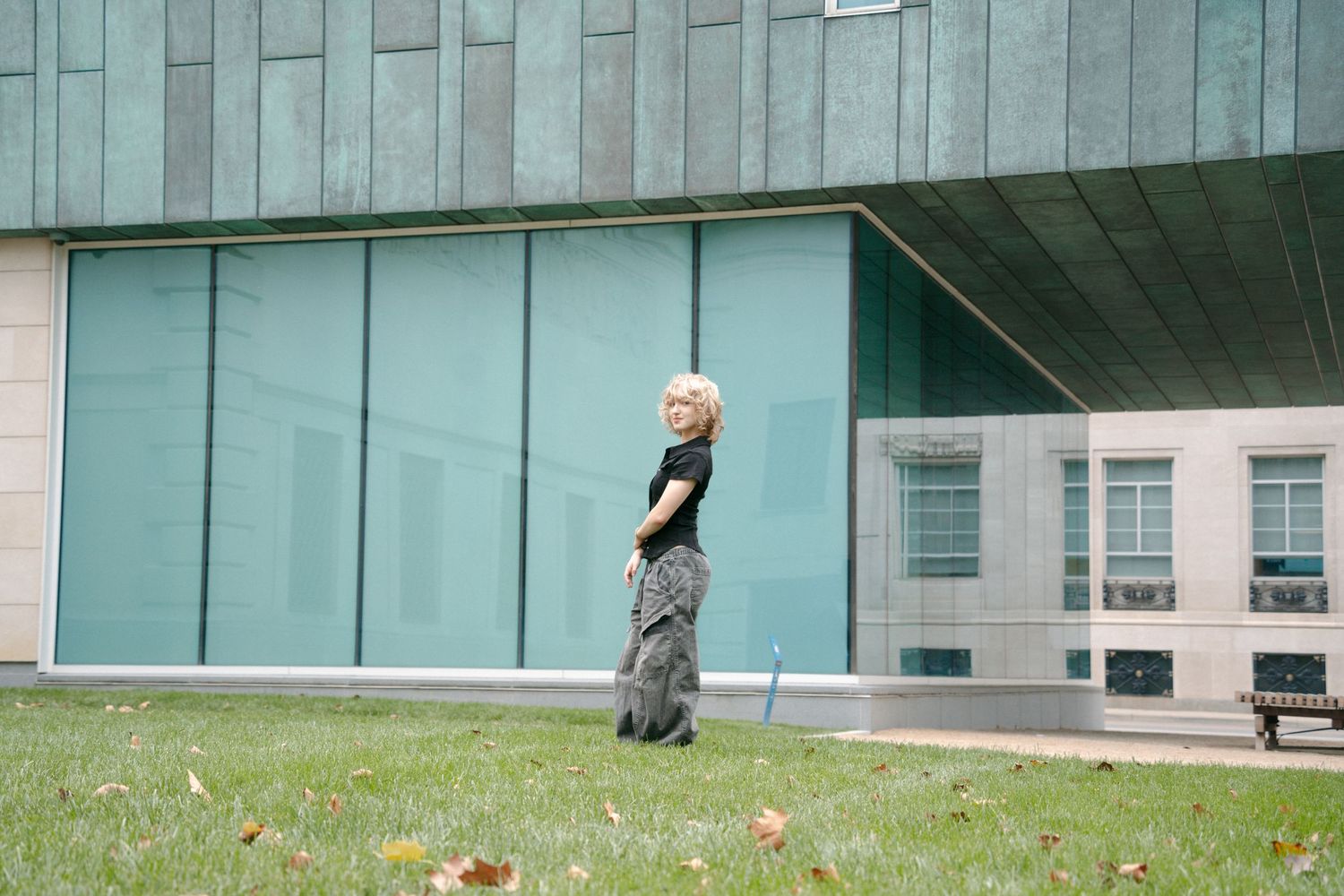 Blonde person in black top and patterned pants standing on grass beside modern glass building exterior.