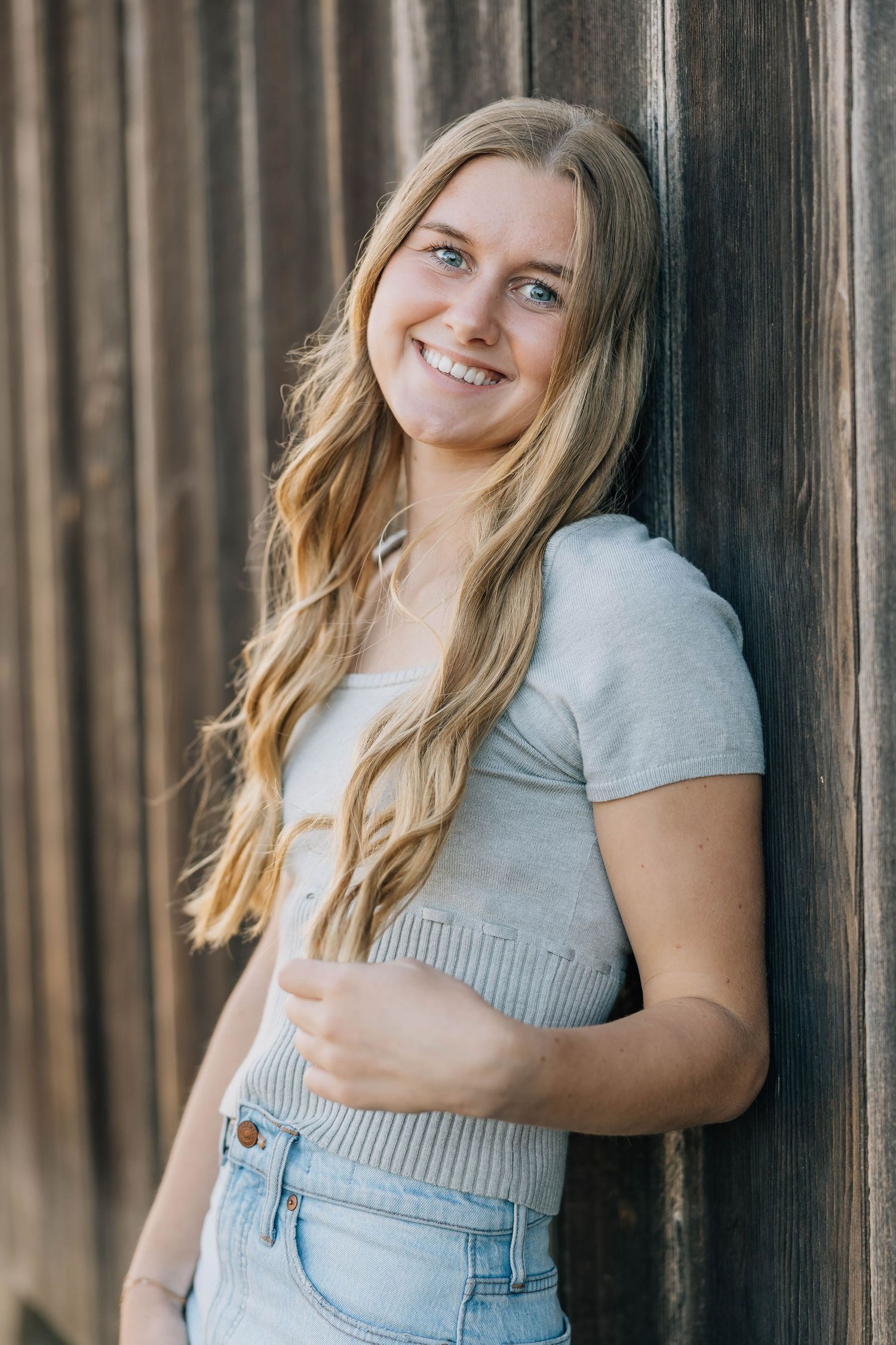 Smiling blonde teenage girl in grey crop top and jeans leans against wooden barn wall.