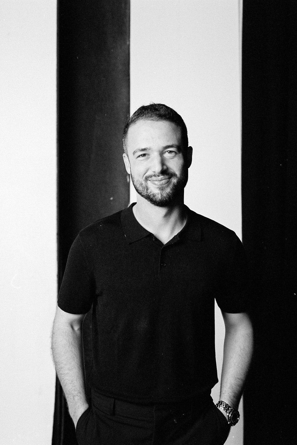 Black and white portrait photograph of a smiling person wearing a dark top outdoors in a forest setting.