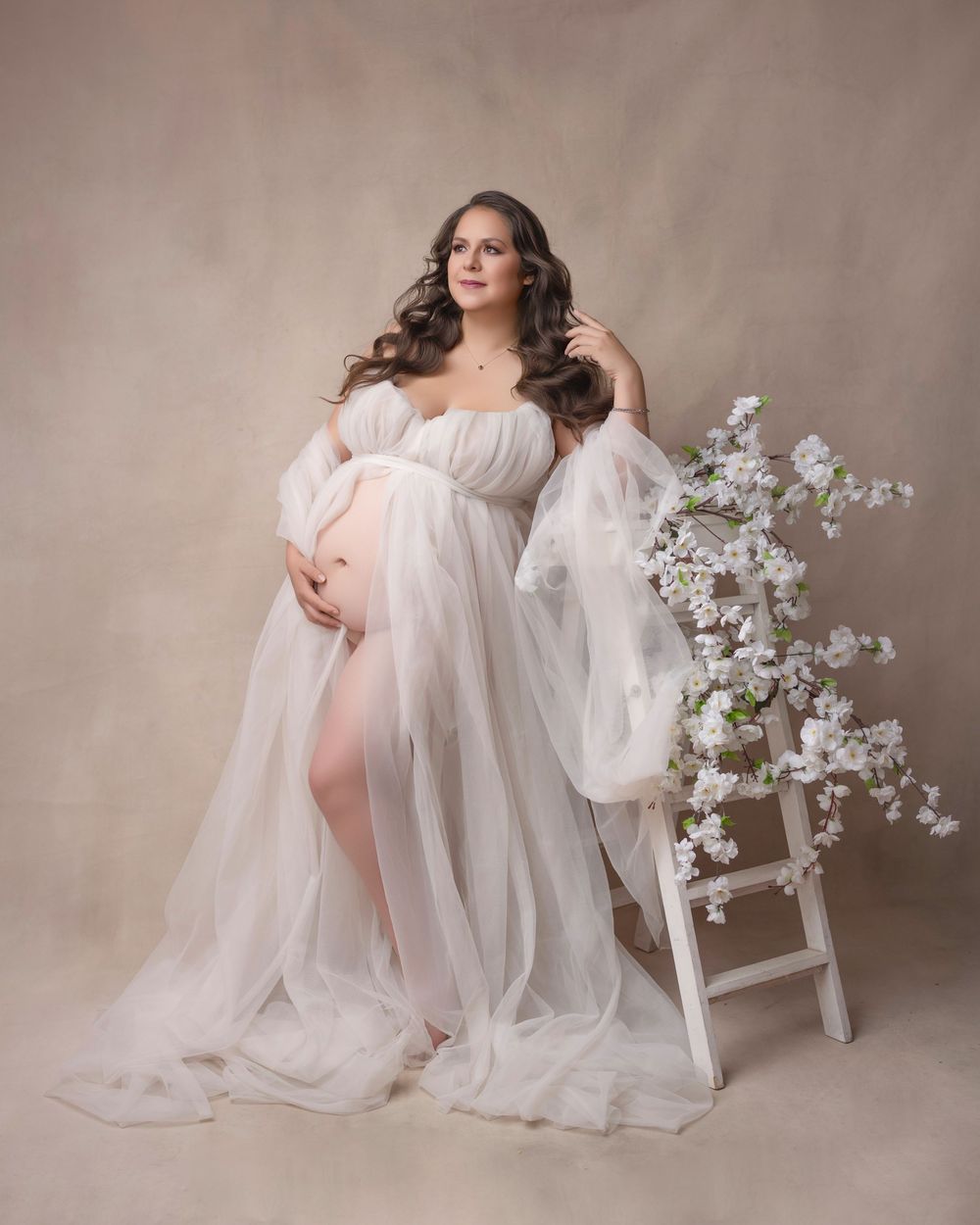 Elegant maternity photo with white sheer gown and cherry blossoms on ladder prop.