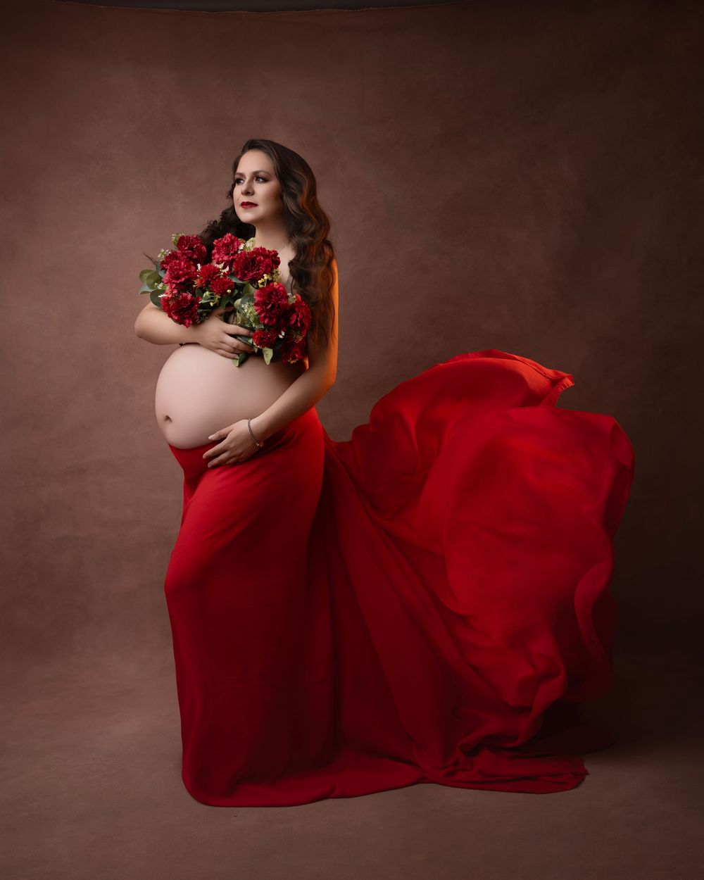 Dramatic maternity photo with red roses and flowing crimson dress against dark background.