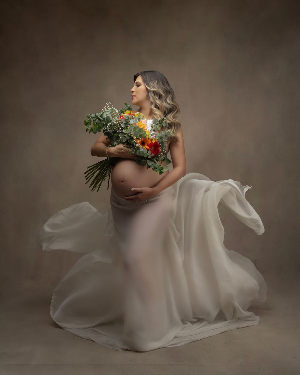 Maternity portrait with flowing white dress and vibrant floral bouquet.