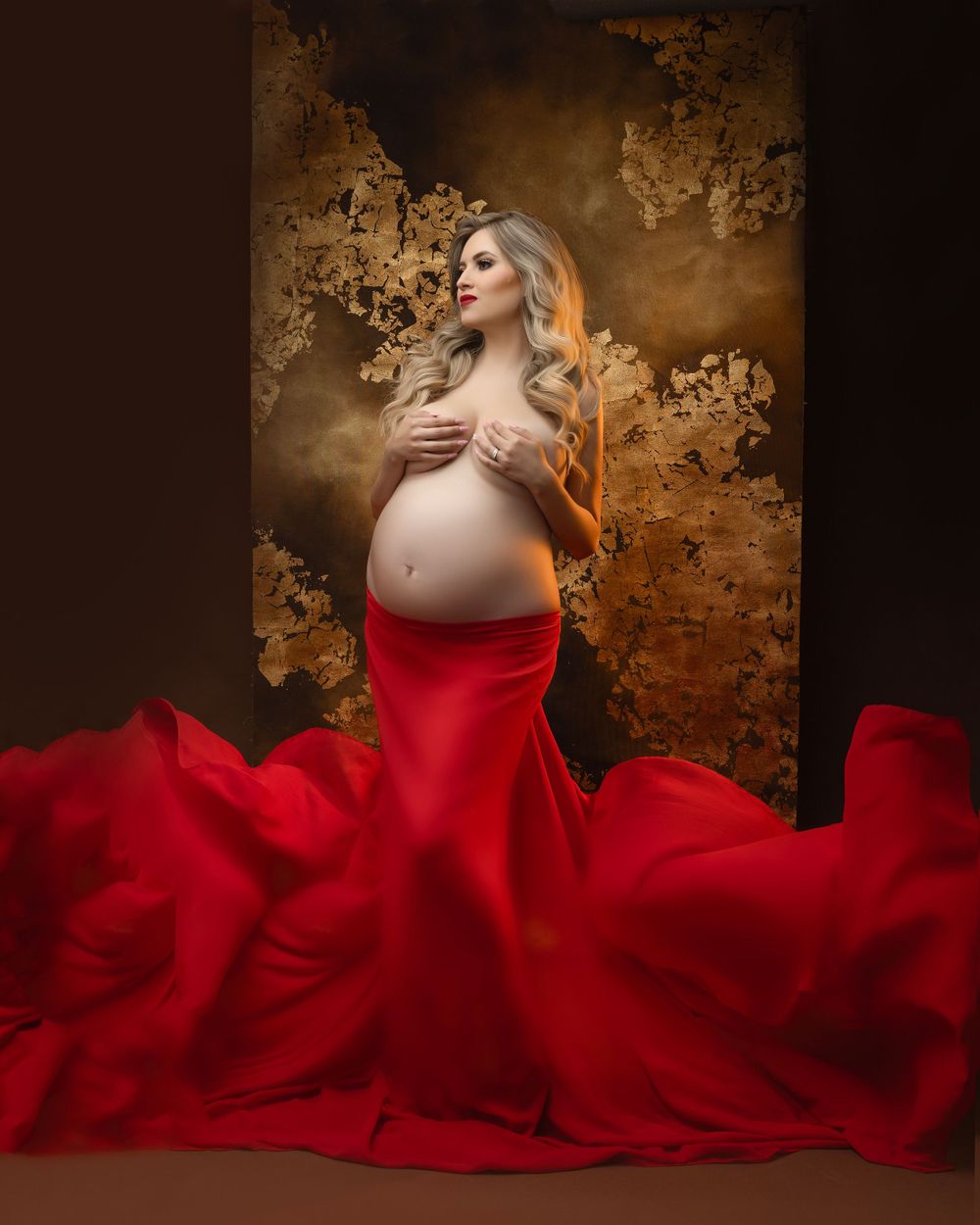 Dramatic maternity portrait featuring flowing red dress against textured golden backdrop.