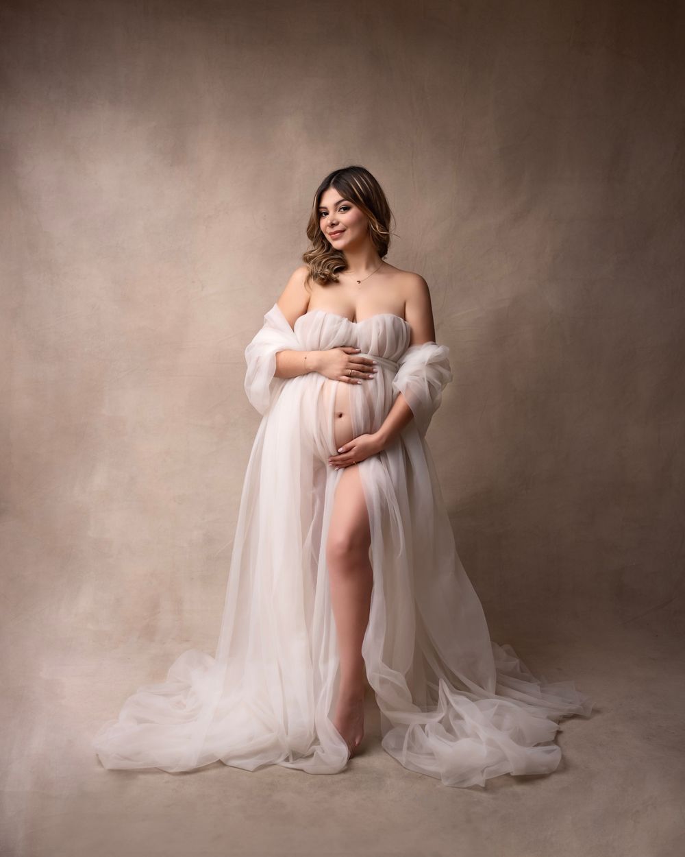 Artistic maternity photo series with soft white tulle fabric draping against a neutral brown background.