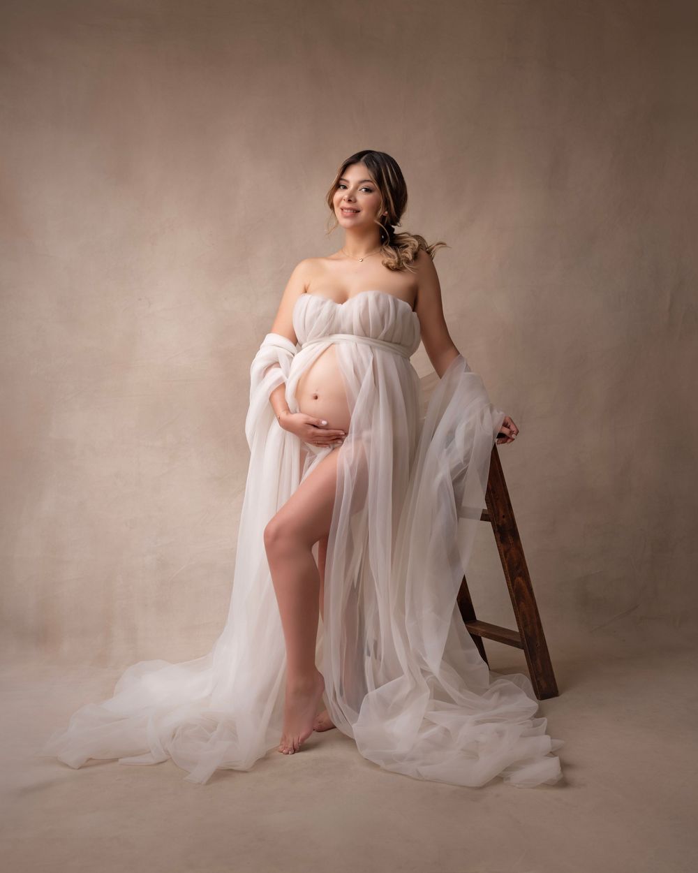 Ethereal maternity portraits featuring delicate white tulle and wooden chair against a brown backdrop.