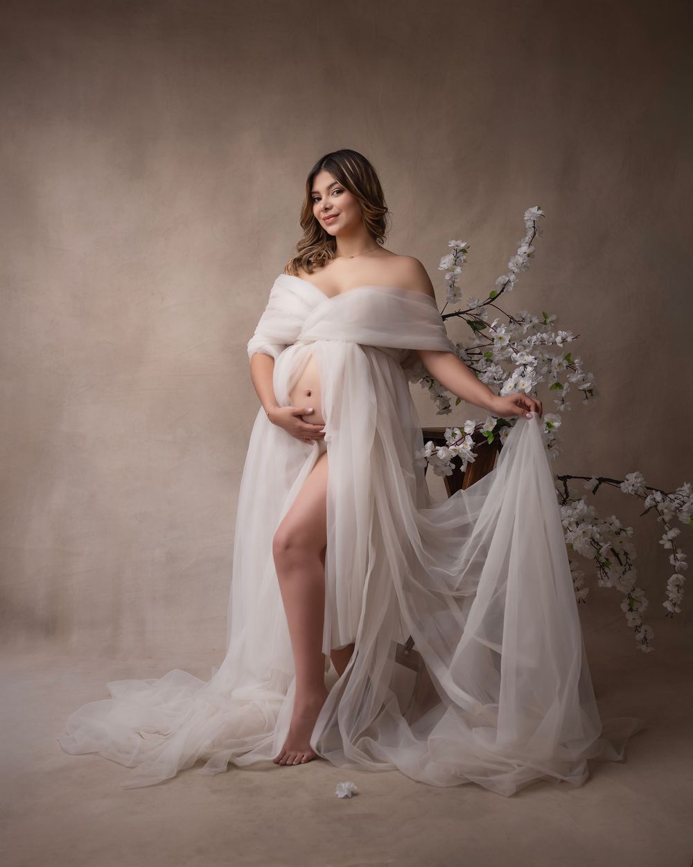 Romantic maternity session with white floral accents and flowing fabric against neutral walls.
