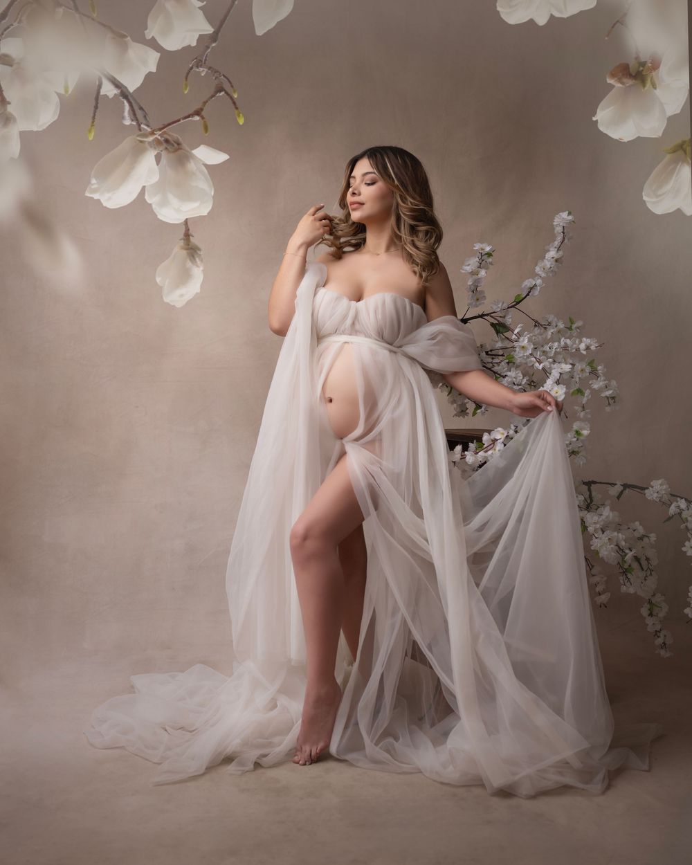 Artistic maternity photos with white cherry blossoms and sheer fabric in a studio setting.