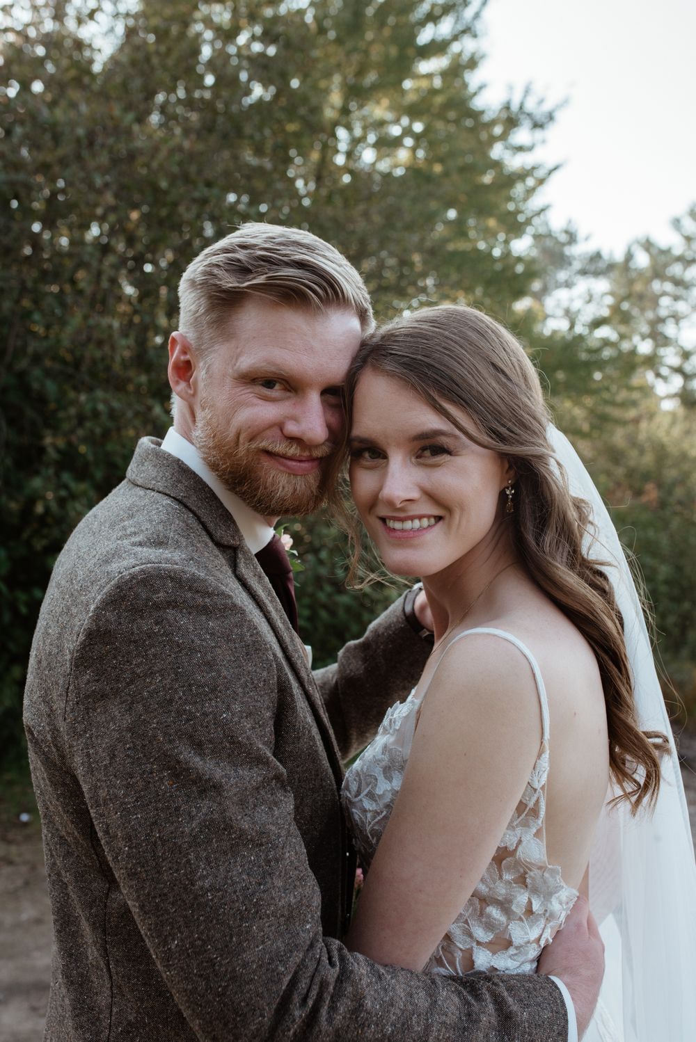 A couple embraces in a wedding photo taken outdoors with evergreen trees visible in the background.