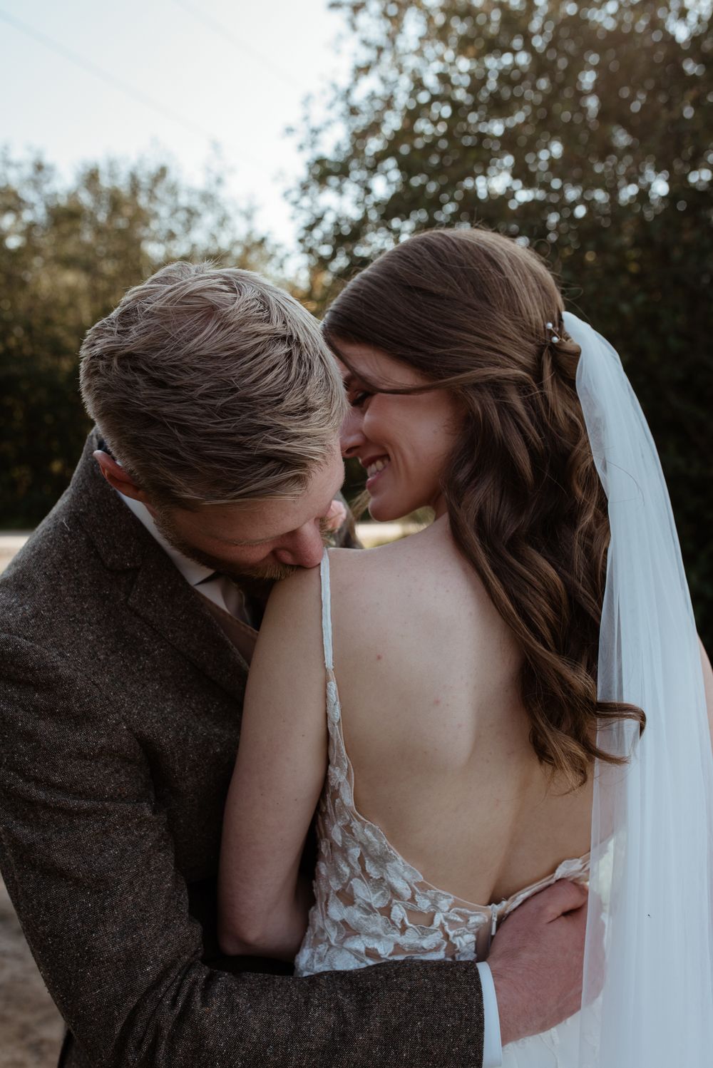 Intimate wedding photo showing couple sharing romantic laughter outdoors with bride in lace backless dress and veil.
