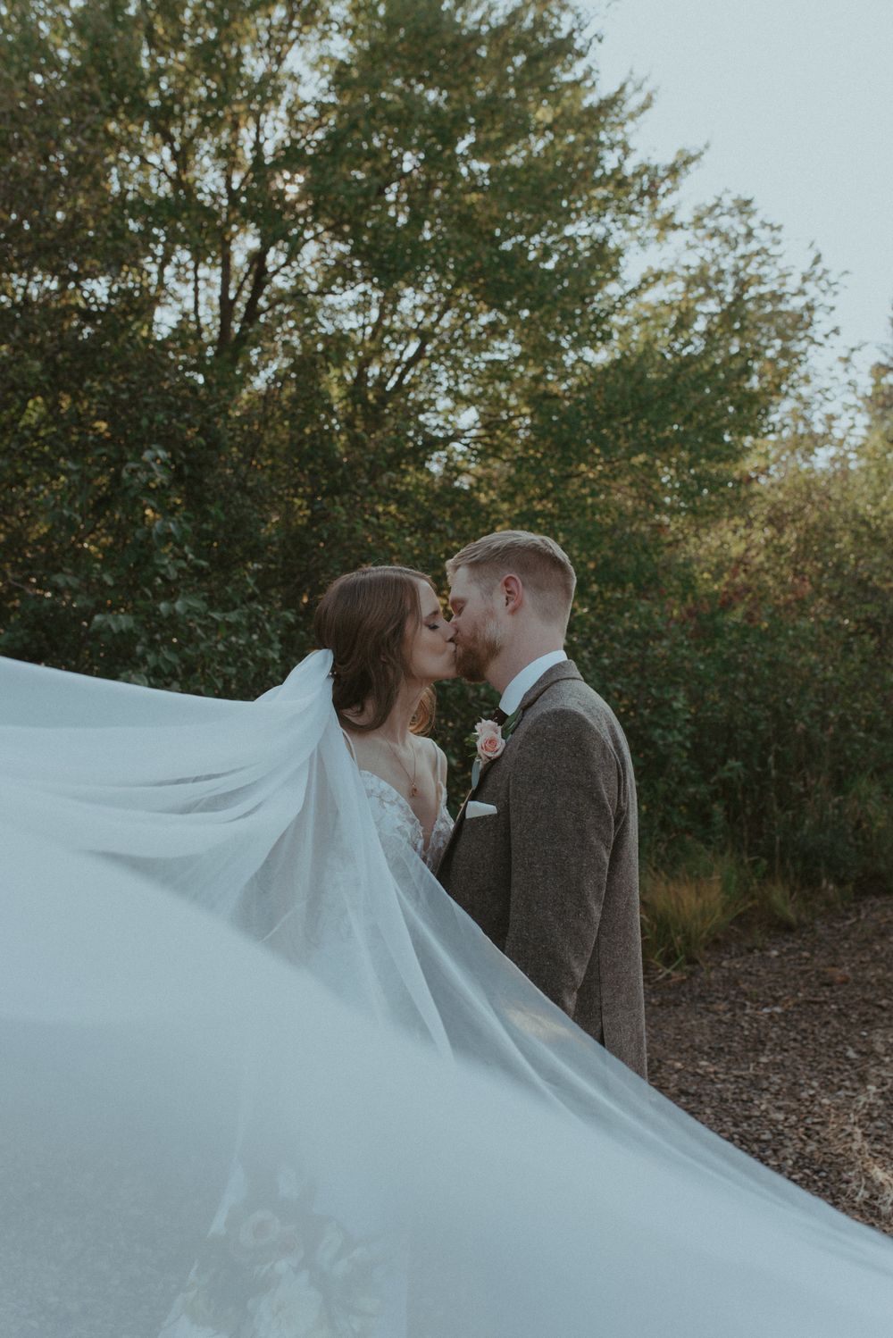 Flowing white veil catches wind behind couple embracing during romantic outdoor wedding moment.