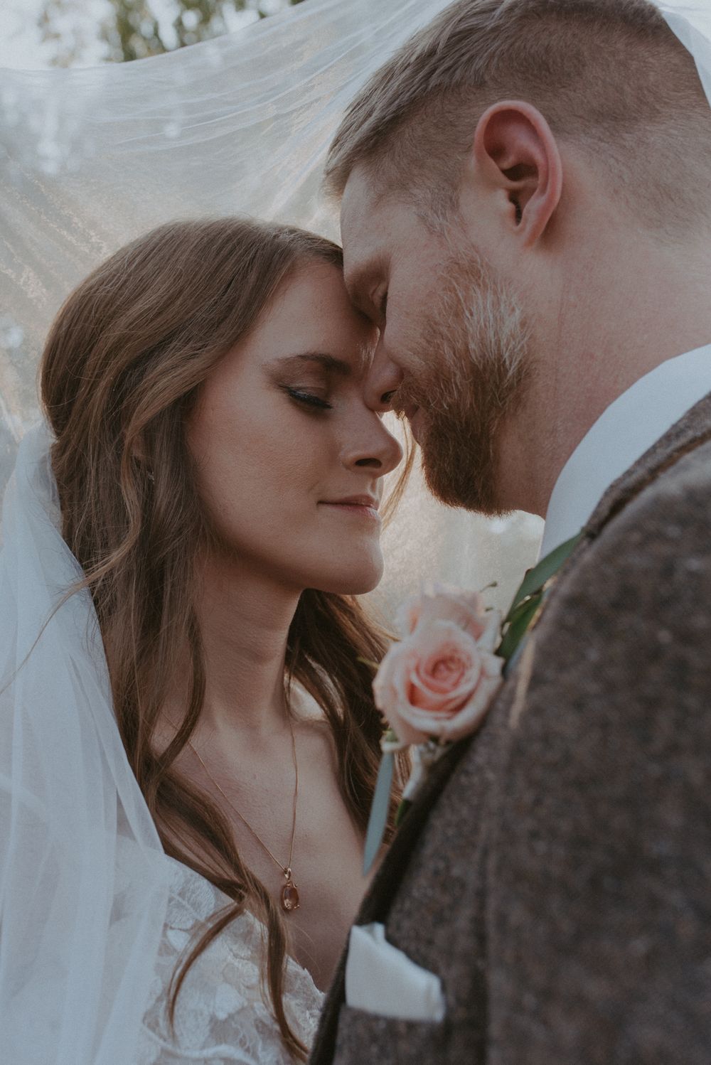A romantic wedding portrait showing a tender moment between newlyweds with a pink rose boutonniere.