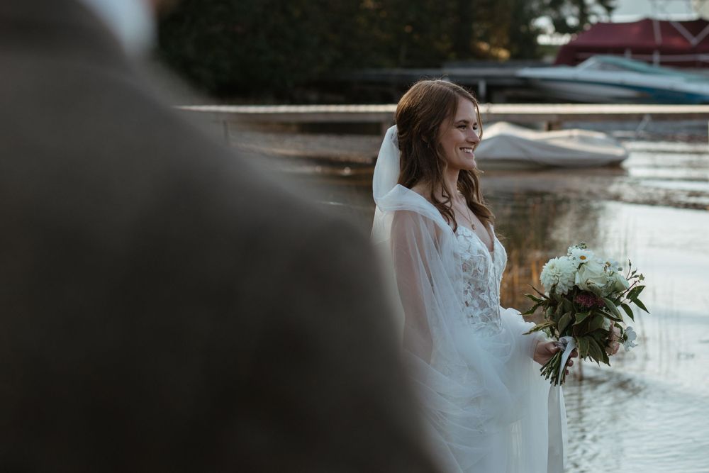 A bride in white dress holds a floral bouquet while standing near water at sunset.