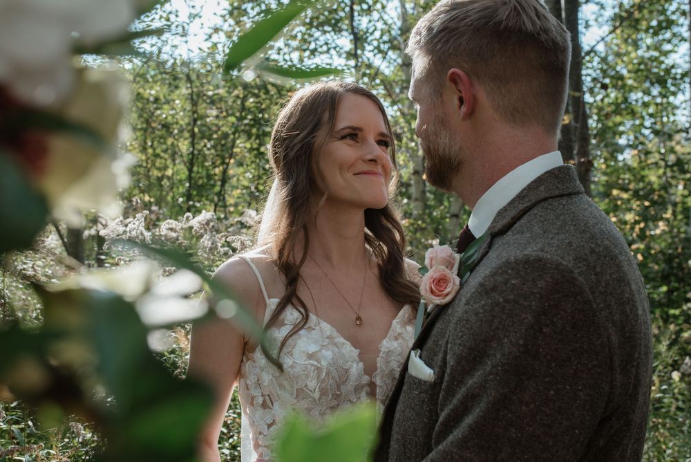 A wedding couple shares an intimate moment in a sun-dappled forest setting with greenery and soft lighting.