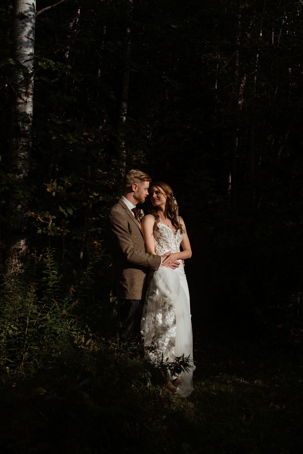 A romantic wedding photo in a dark moody forest setting with soft light illuminating a couple in elegant attire.