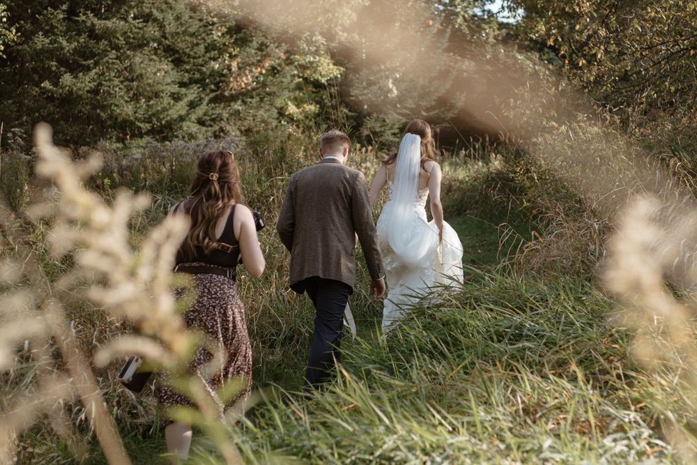 Wedding photographer walks down a sunlit garden path through tall grasses and wildflowers on a summer day.