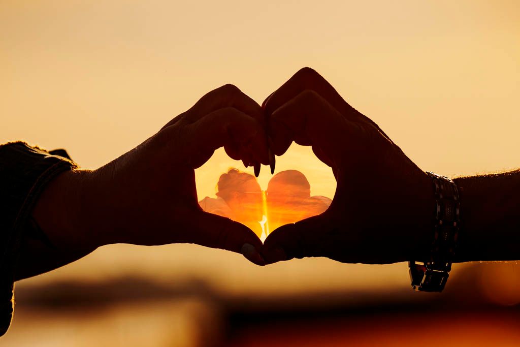 Silhouette of hands forming a heart shape against a sunset sky.