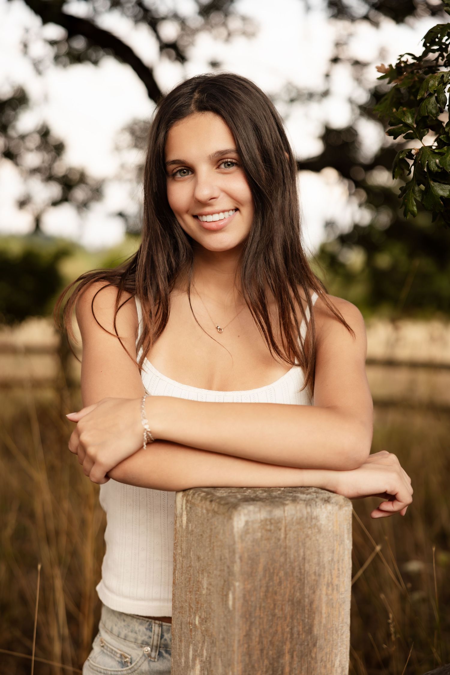 A young person in a white tank top leans against a wooden post in a sunlit outdoor rural setting during golden hour.